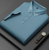 Camicia polo blu piegata su sfondo nero, con dettagli a righe sul colletto e tre bottoni. Orologio da polso elegante con cinturino in pelle accanto.